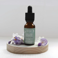 Douceur Florale – Parfum de linge léfé, flacon compte-gouttes ambré avec étiquette verte, entouré de fleurs de violette et blanches